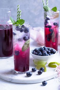 two glasses filled with blueberries and limeade on a marble tray next to some minty garnishes