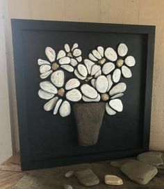 a vase with white flowers in it sitting next to some rocks on the ground and a black frame