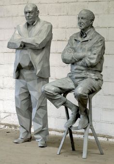 two statues of men sitting on stools in front of a white brick wall, one with his arms crossed