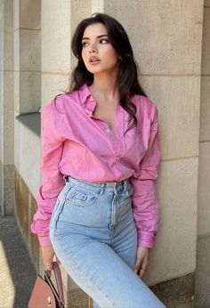 a woman leaning against a wall with her hand on her hip wearing jeans and a pink shirt