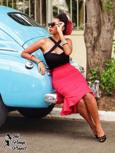 a woman sitting on the back of a blue car talking on a cell phone while wearing a black top and pink skirt