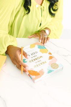 a woman sitting at a table with a book and pencil in her hand, writing on it