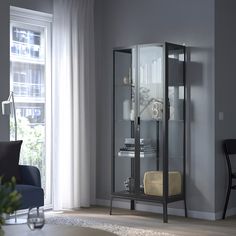 a tall glass cabinet in the corner of a living room next to a chair and window