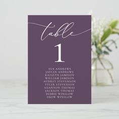 a purple table number card with white calligraphy on it and a vase full of flowers in the background