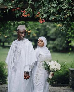 a man and woman dressed in white holding hands under a tree with flowers on it