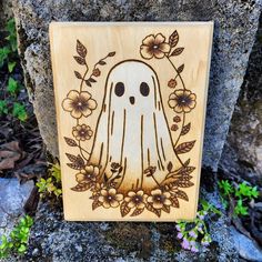 a carved wooden plaque with a ghost surrounded by flowers and leaves on top of a rock