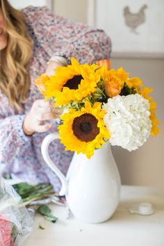 a woman arranging sunflowers in a white pitcher