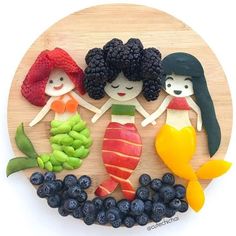 three little mermaids made out of fruit on a wooden platter with grapes, raspberries and blueberries