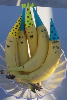 four bananas with faces drawn on them sitting in a glass bowl, wearing party hats