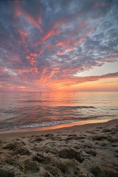 the sun is setting over the ocean with clouds in the sky and sand on the beach