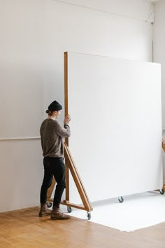 a man standing next to an easel on top of a hard wood floor in front of a white wall