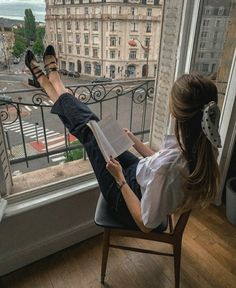 a woman sitting in a chair reading a book on top of a window sill