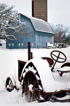 an old tractor is covered in snow near a blue barn and silo on a snowy day