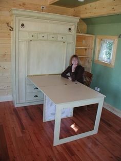 a woman sitting at a table in a room with wooden floors and walls, she has her hands on her chin