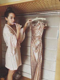 a woman brushing her teeth while standing next to a dress on a hanger in a closet