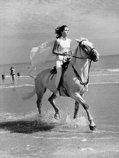 a woman riding on the back of a white horse down a beach next to the ocean