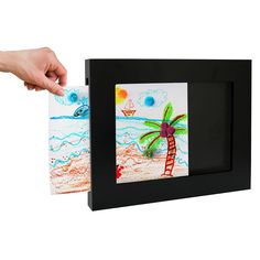 a hand is holding an art project in front of a picture frame with a palm tree on it