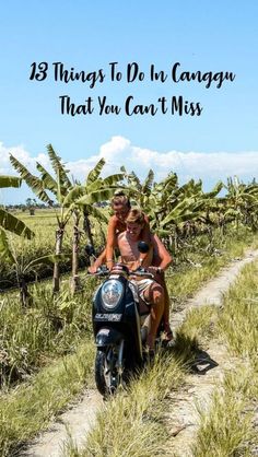 two people riding on the back of a motorcycle down a dirt road with palm trees in the background