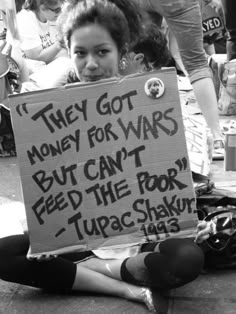 a woman sitting on the ground holding a sign that says they got money for wars but can't feed the poor tupac shaka