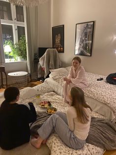 three people sitting on the floor in a bedroom with two beds and one is eating food