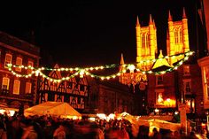 people are walking around an outdoor market with christmas lights strung across the street and buildings in the background