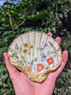 a hand holding a flowered fan in front of some bushes