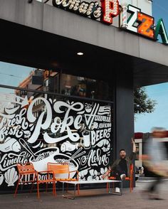 a man sitting on an orange chair in front of a pizza shop with graffiti all over it