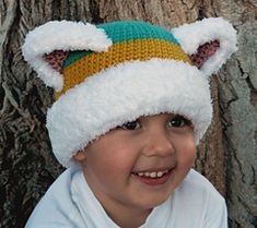 a young boy wearing a knitted hat with ears and eyes, standing next to a tree