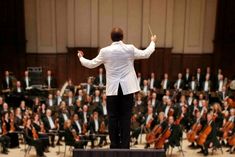 conductor in white jacket and black pants standing at the front of an orchestra with his arms outstretched