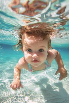 A smiling baby swimming underwater with bubbles, wearing a light blue swimsuit with floral details, surrounded by clear turquoise water.