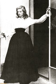 a woman in a long black skirt is standing by an open door with her hand on the handle