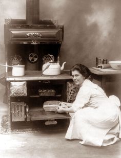 an old black and white photo of a woman sitting in front of a stove holding a loaf of bread