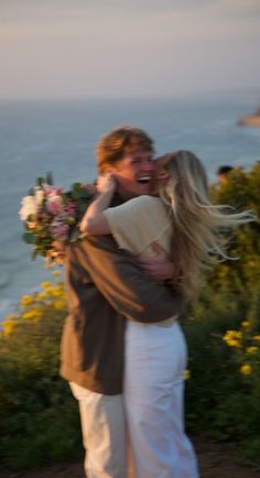 a man and woman embracing each other in front of the ocean with wildflowers