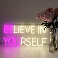 a vase filled with pink flowers next to a neon sign that says believe in yourself