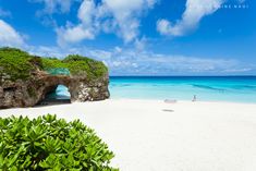 a sandy beach next to the ocean under a blue sky with white clouds and green vegetation