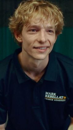 a young man with blonde hair wearing a black polo shirt and smiling at the camera