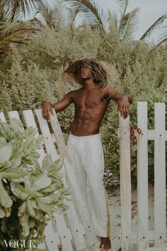 a man with no shirt standing next to a white picket fence and holding a straw hat on his head