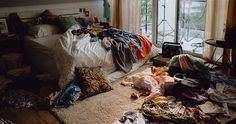 an unmade bed with lots of clothes on the floor in front of a window