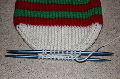 two knitting needles are laying next to a knitted hat with red, white and green stripes