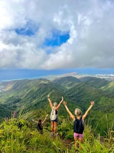 two women standing on top of a lush green hillside with their arms in the air