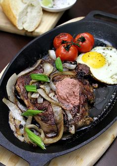 an iron skillet with steak, onions and tomatoes on it next to sliced bread