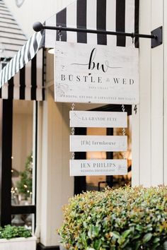 a sign hanging from the side of a building next to bushes and potted plants