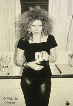 a woman with curly hair standing in front of a kitchen counter holding a card and looking at the camera