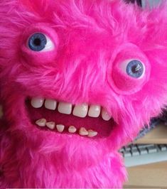 a pink stuffed animal with big blue eyes and teeth is sitting on a desk next to a computer