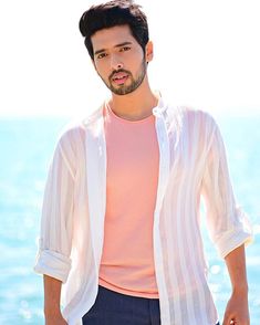 a man standing in front of the ocean wearing a pink shirt and blue pants with his hands in his pockets