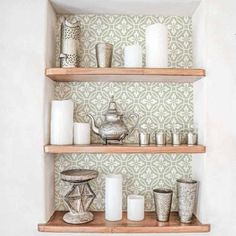 shelves with candles, teapots and cups on them in front of a wallpapered background