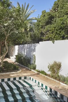 an empty pool in the middle of a garden with trees and bushes around it, next to a white fence