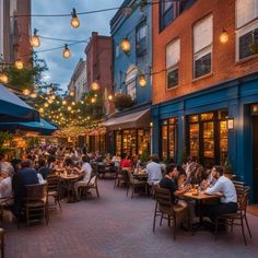 people are sitting at tables in an outdoor dining area with lights strung from the buildings