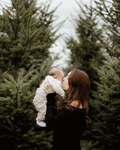 a woman holding a baby in her arms and kissing it's face with trees behind her