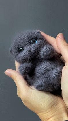 a hand holding a small gray stuffed animal with blue eyes and one eye open, in front of a grey background